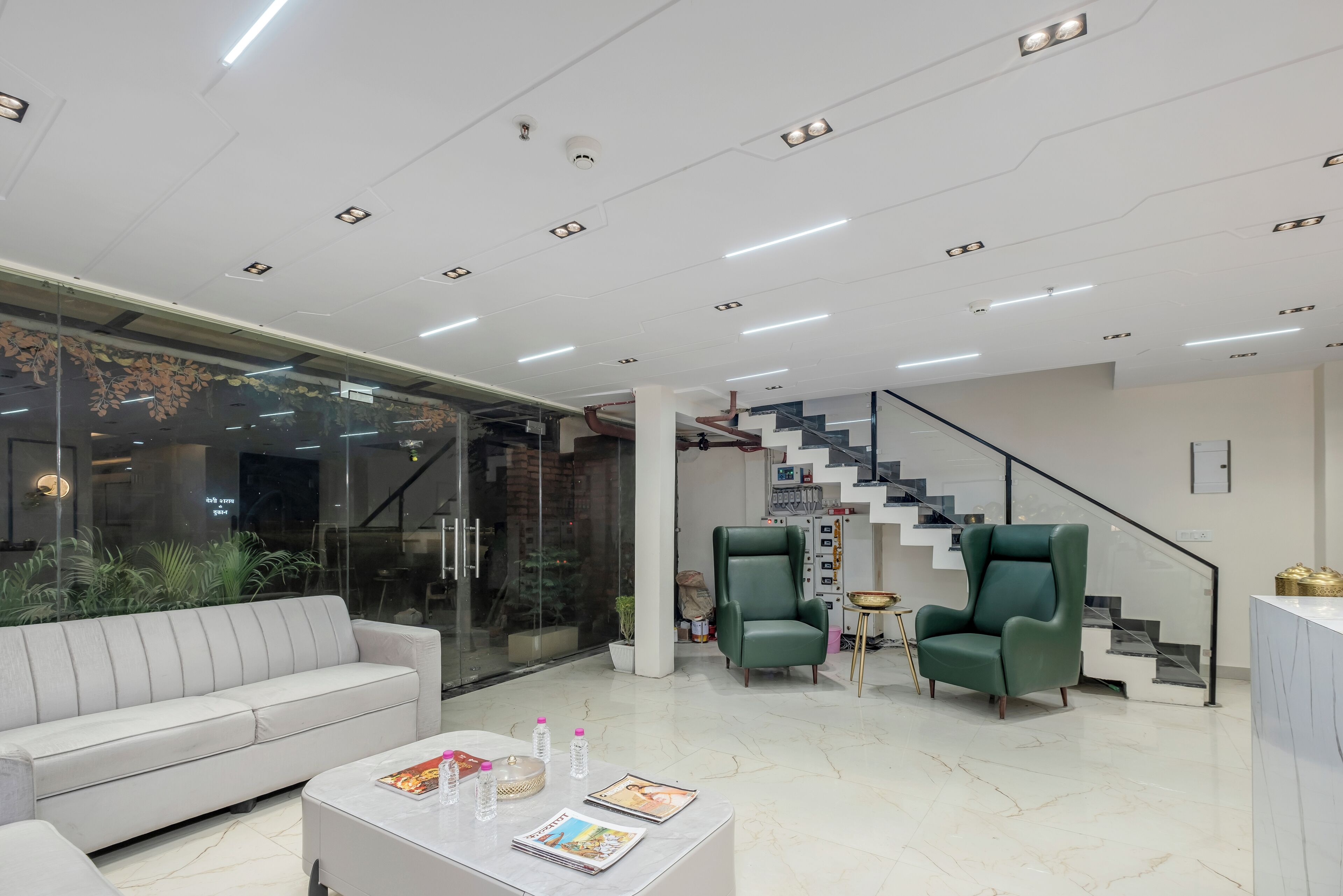 A modern interior with a white sofa, a coffee table with magazines, two green armchairs, and a staircase with a black railing.