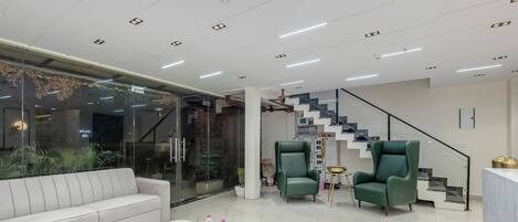 A modern interior with a white sofa, a coffee table with magazines, two green armchairs, and a staircase with a black railing.