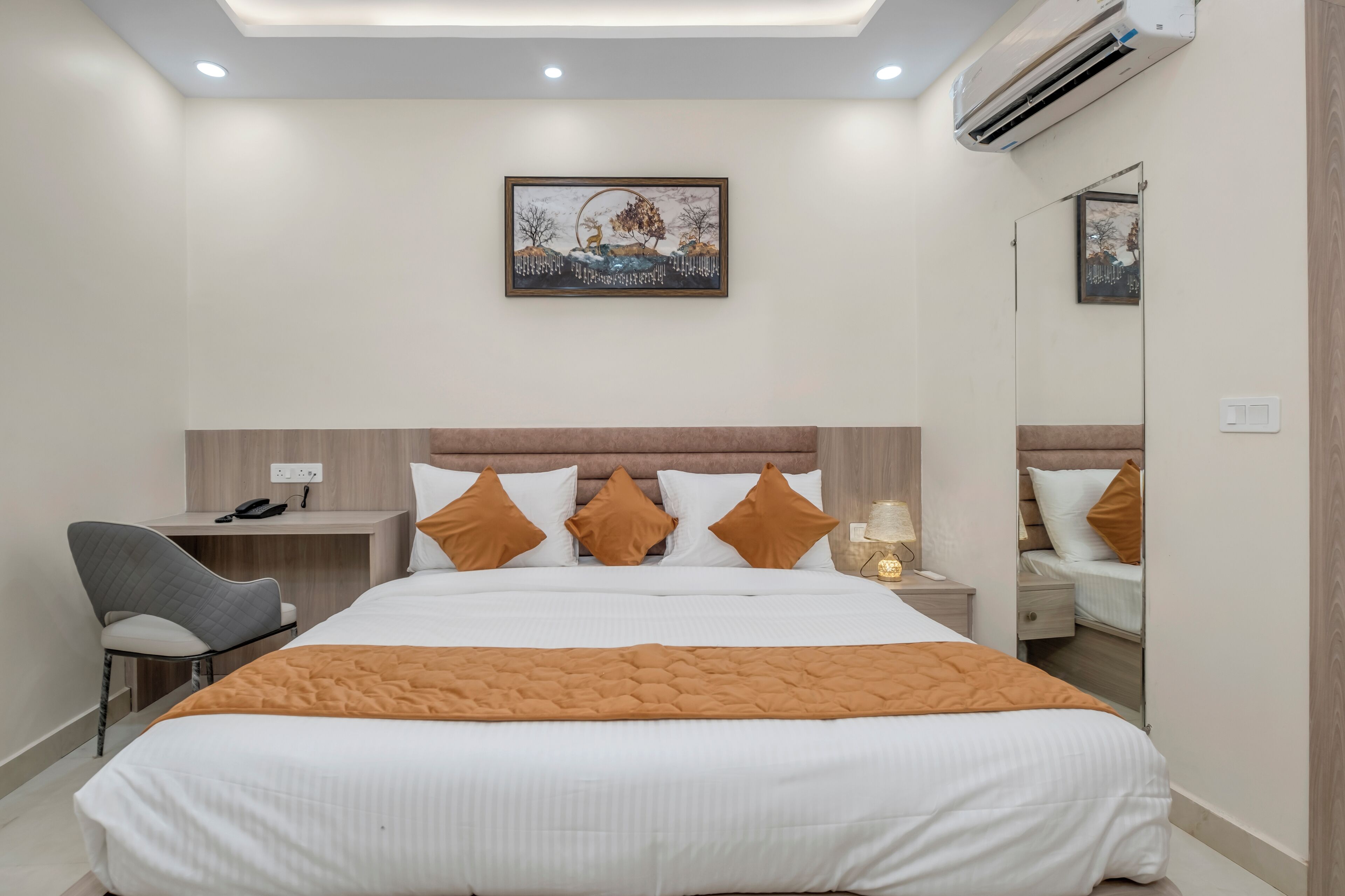 A modern hotel room with a large bed, a desk, and a chair. There is a painting on the wall and an air conditioning unit above the bed.