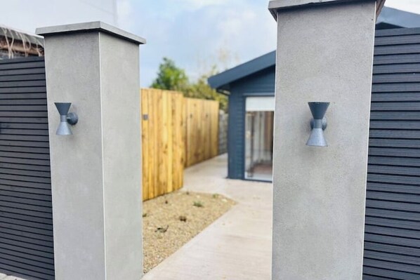 Une entrée extérieure moderne, avec des piliers en béton et des éléments métalliques, qui mène à une clôture en bois et à un bâtiment à la façade sombre.
