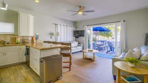 A modern kitchen with white cabinets, a central island, and a built-in oven. There is a dining area with a sofa and a small table. - Coastal Haven (San Diego)