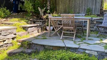 Un patio con una mesa de piedra y sillas de madera, rodeado por un muro de piedra y vegetación.