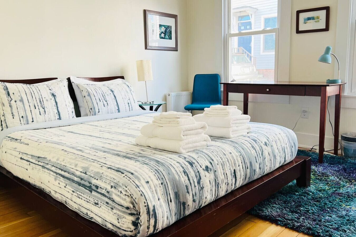 Un lit bien fait, recouvert d’une couverture à rayures, une pile de serviettes blanches, une chaise bleue, un bureau avec une lampe et une fenêtre donnant sur un immeuble.