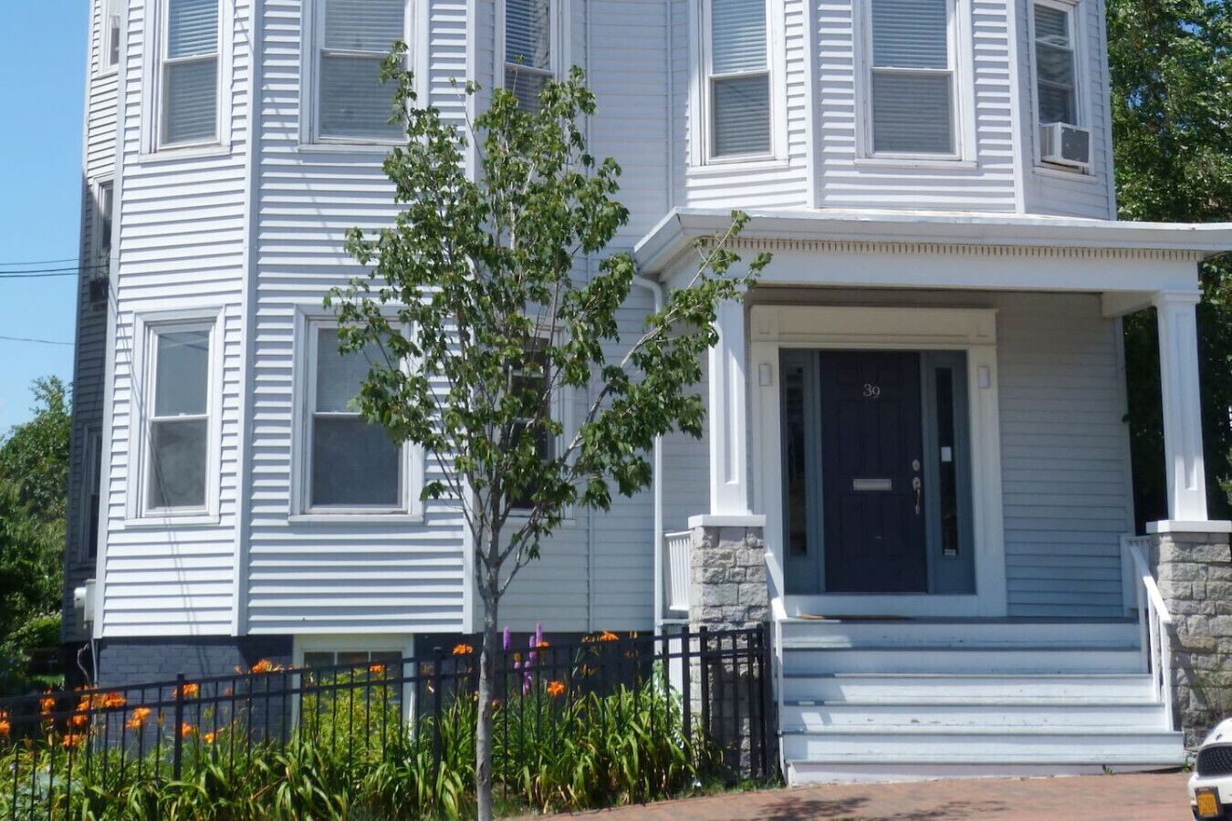 Une maison blanche à deux étages, avec une porte d’entrée bleu foncé, un petit porche et un arbre devant.