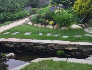 Un jardín con un puente de piedra sobre un pequeño arroyo, rodeado de diversas plantas y flores.