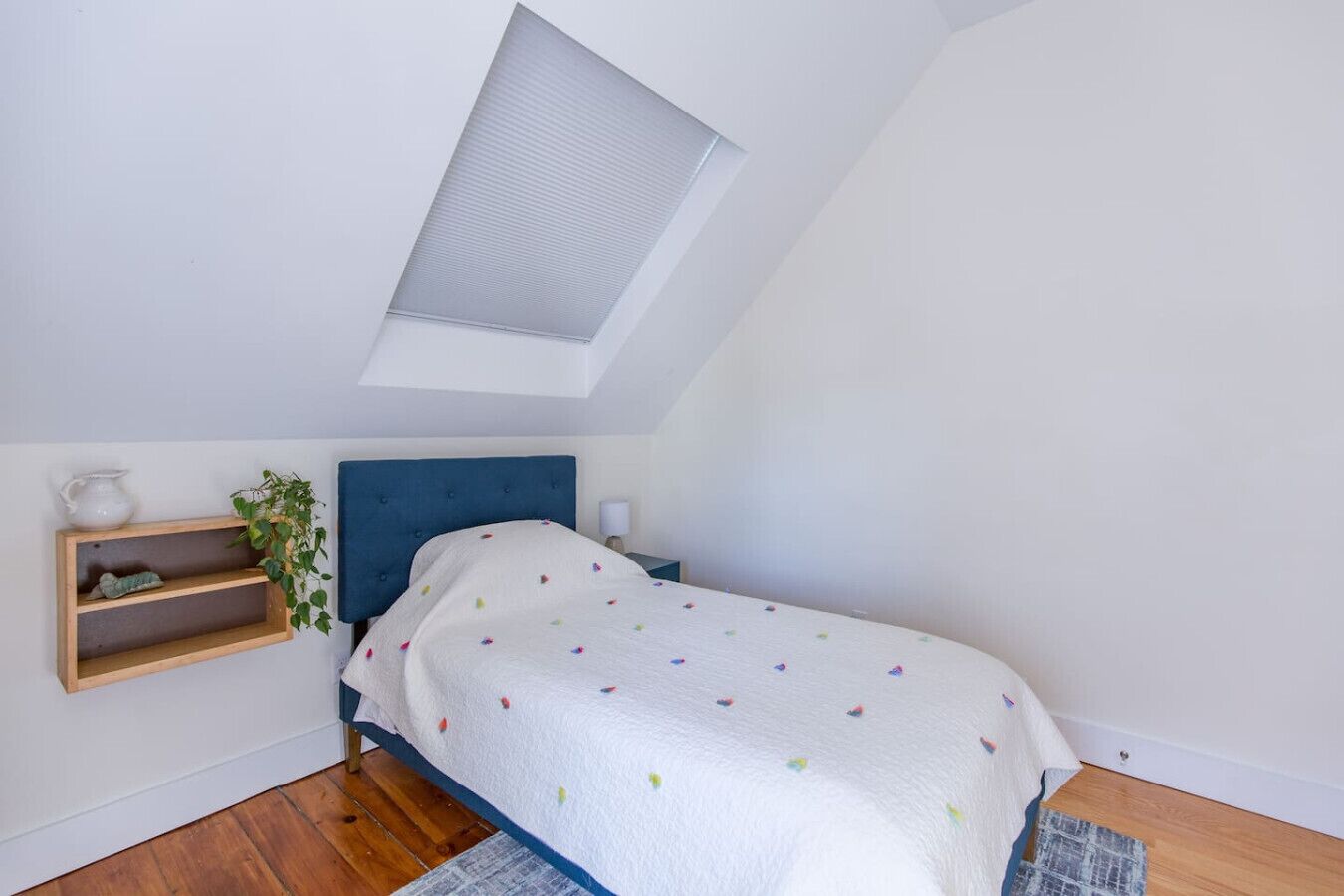 Ein Schlafzimmer mit schrägen Wänden, einem Bett mit weißer Decke, einem blauen Kopfteil, einem Holzregal mit einer Pflanze und einem Oberlicht.