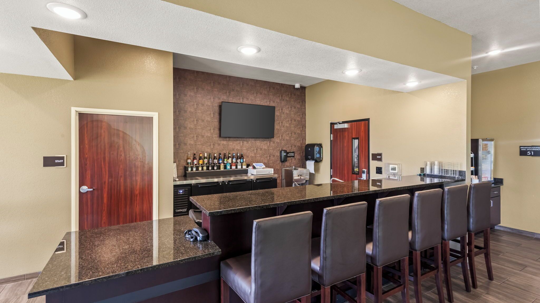 Un bar de hotel con una barra alargada, varias sillas, un televisor de pantalla plana y un minibar.