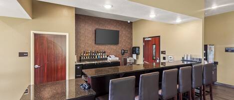 Un bar de hotel con una barra alargada, varias sillas, un televisor de pantalla plana y un minibar.