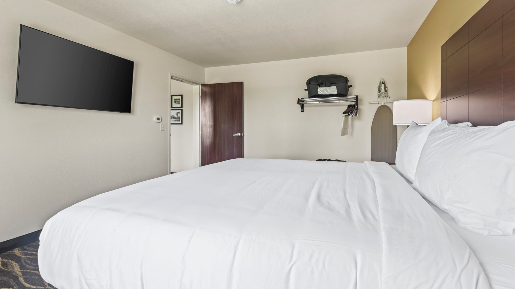 A hotel room with a large bed, a flat-screen TV mounted on the wall, a coat rack with a jacket, and a door leading to another room.