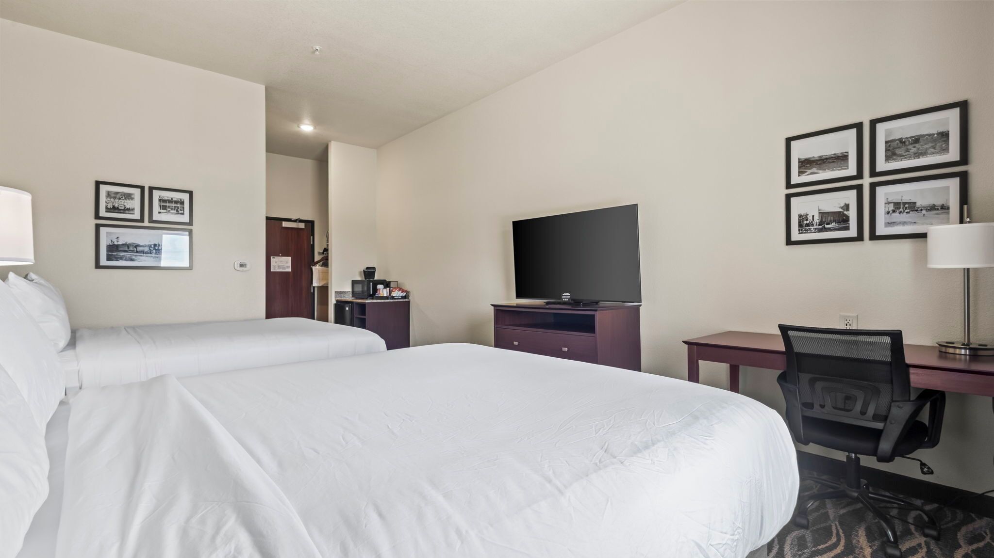 Habitación de hotel con dos camas, un escritorio con computadora, televisor y cuadros en la pared.