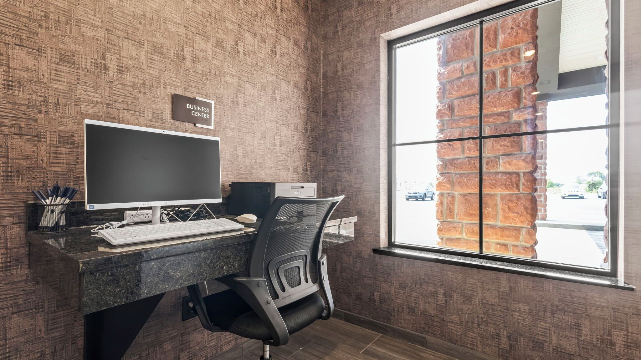 Ein moderner Arbeitsraum mit Ziegelwand, großem Fenster, Schreibtisch mit Computer und Stuhl.