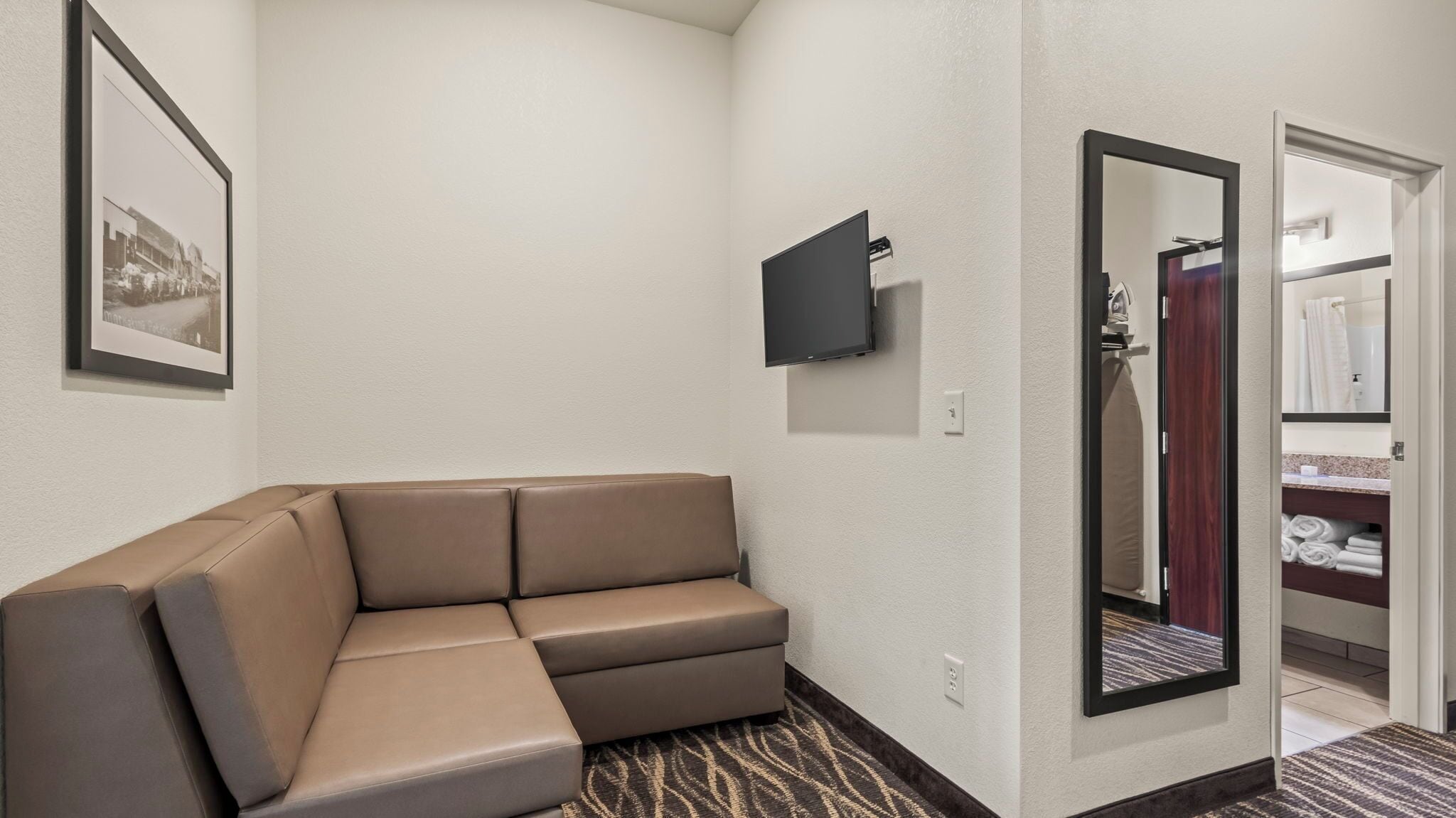 A hotel room with a beige sofa, a flat-screen TV mounted on the wall, and a mirror reflecting a bathroom with towels and a shower.