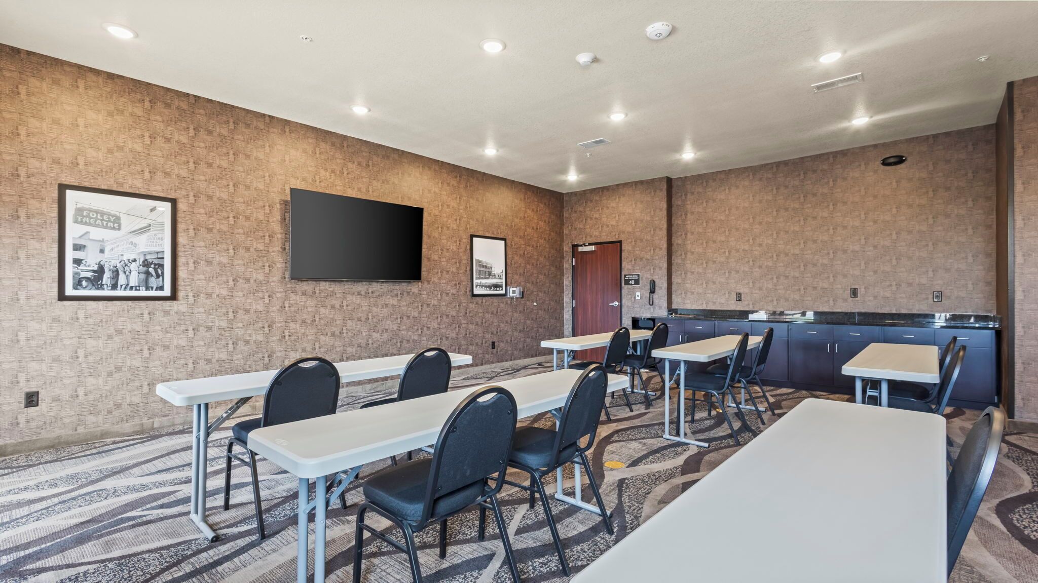 A conference room with tables and chairs, a flat-screen TV mounted on the wall, and a kitchenette area.