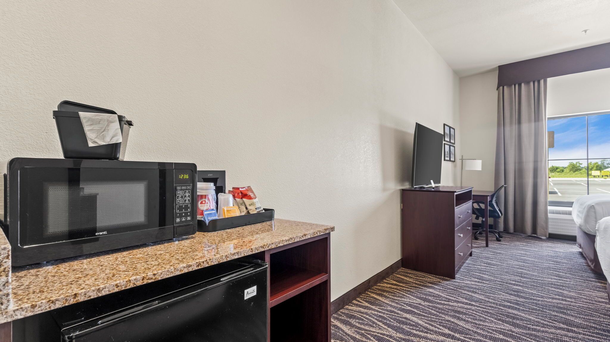 Habitación de hotel con kitchenette, televisión de pantalla plana, escritorio y ventana grande con vista al estacionamiento.