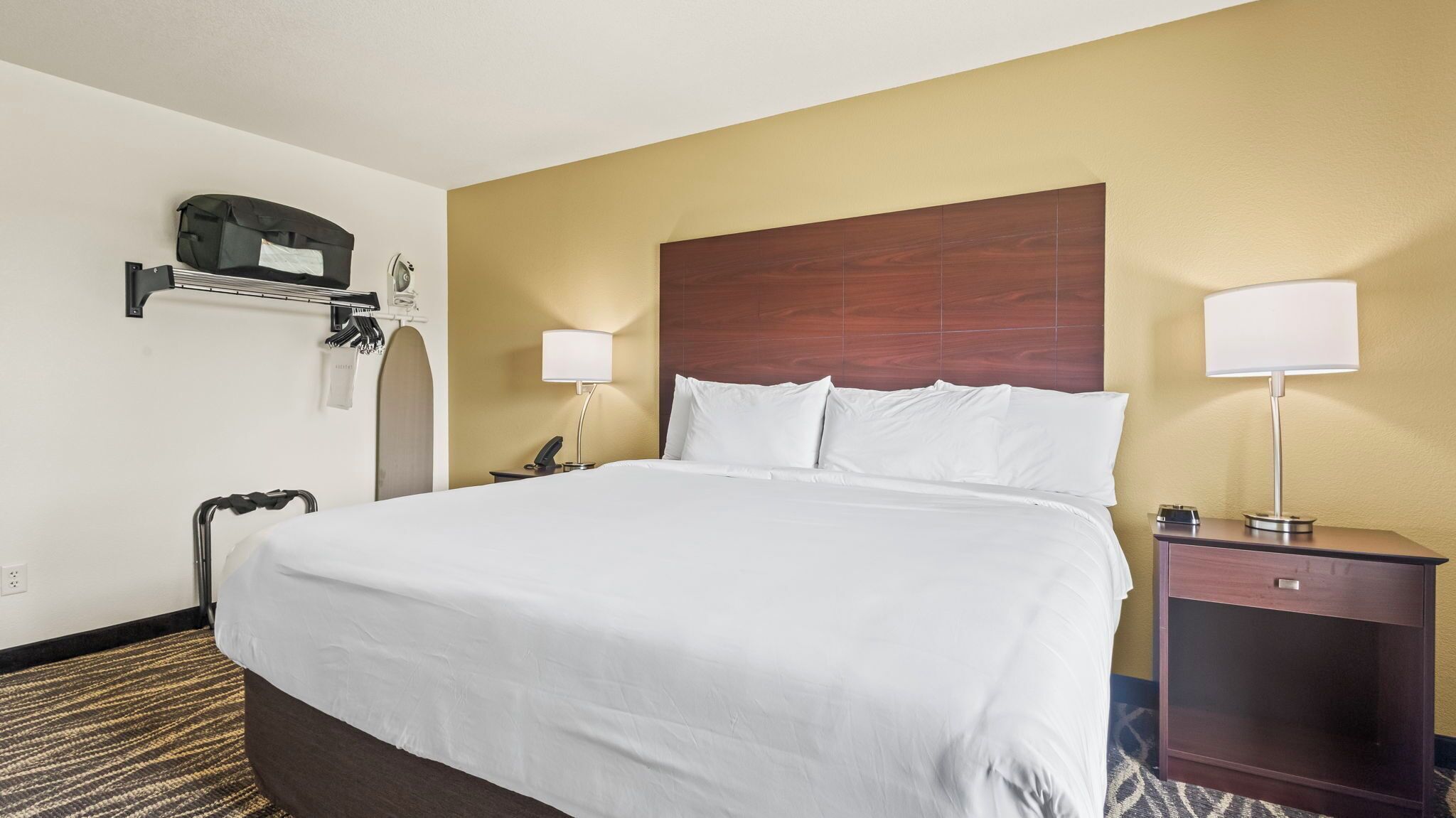 A hotel room with a large bed, a nightstand with a lamp, a desk with a chair, and a coat rack with a bag.