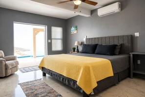 Un dormitorio con cama, ventilador de techo, unidad de aire acondicionado, mesita de noche, lámpara, silla, alfombra y vistas al exterior.