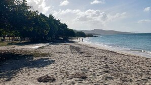 Ein sandiger Strand mit sanften Wellen, wolkenlosem Himmel und Hügeln in der Ferne.