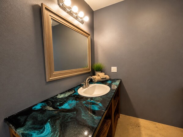 A bathroom with a marble countertop, a sink, a mirror with lights, a potted plant, and a towel rack.