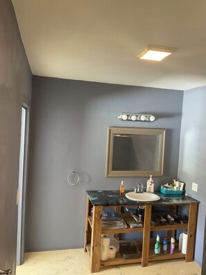 Une salle de bain avec un comptoir en marbre, un lavabo, un miroir et une étagère en bois où sont rangés les articles de toilette.