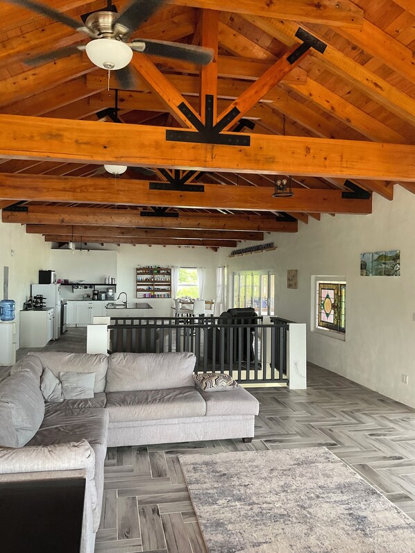A spacious living area with a large gray sectional sofa, a herringbone-patterned wooden floor, and exposed wooden beams. A kitchen with white cabinets and a dining area with a black railing are visible in the background. - New 4-bedroom house overlooking the ocean with beautiful view and ocean sound (Christiansted)
