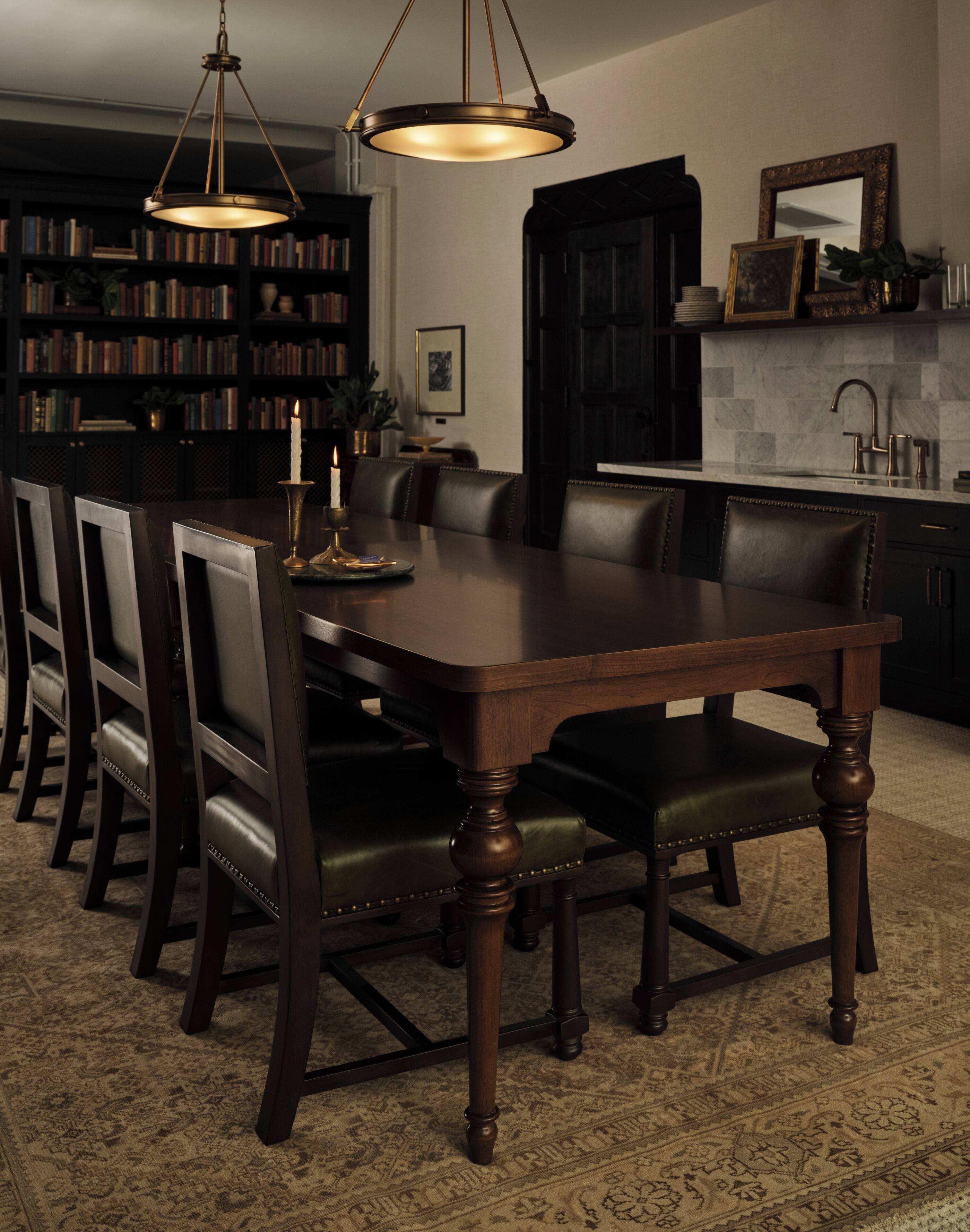 Un coin repas avec une table en bois foncé entourée de chaises, un lustre, une bibliothèque et un espace cuisine comprenant un évier et un miroir.