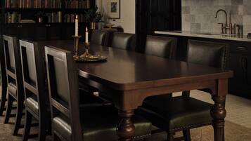 Un coin repas avec une table en bois foncé entourée de chaises, un lustre, une bibliothèque et un espace cuisine comprenant un évier et un miroir.
