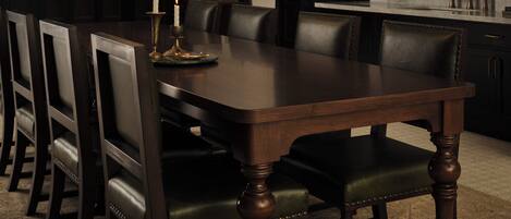 Comedor con mesa de madera oscura rodeada de sillas, una araña, una estantería y una zona de cocina con un lavamanos y un espejo.