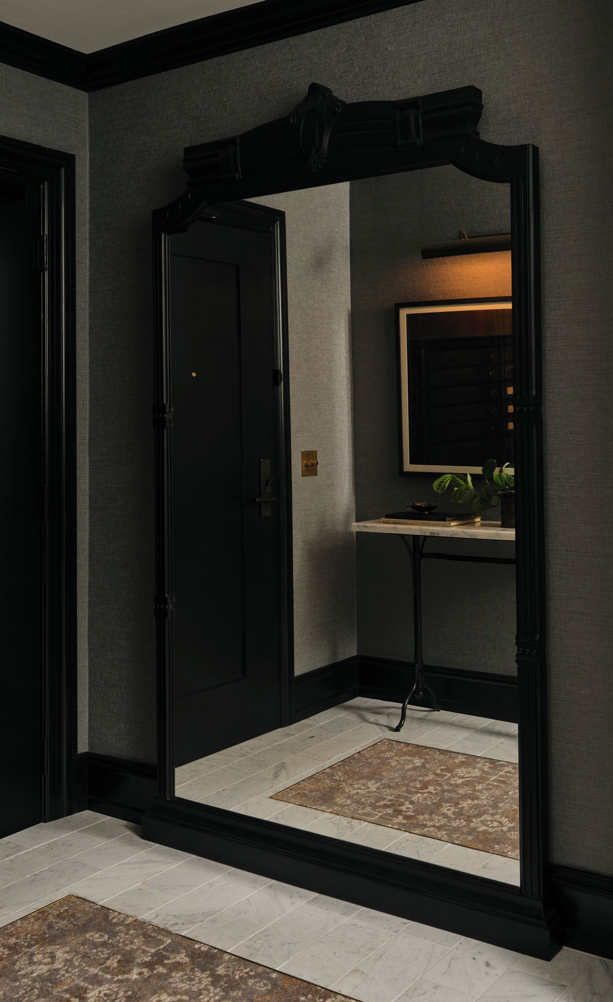 A hallway with a black door, a mirror reflecting a room with a sink and a plant, and a patterned rug on the floor.