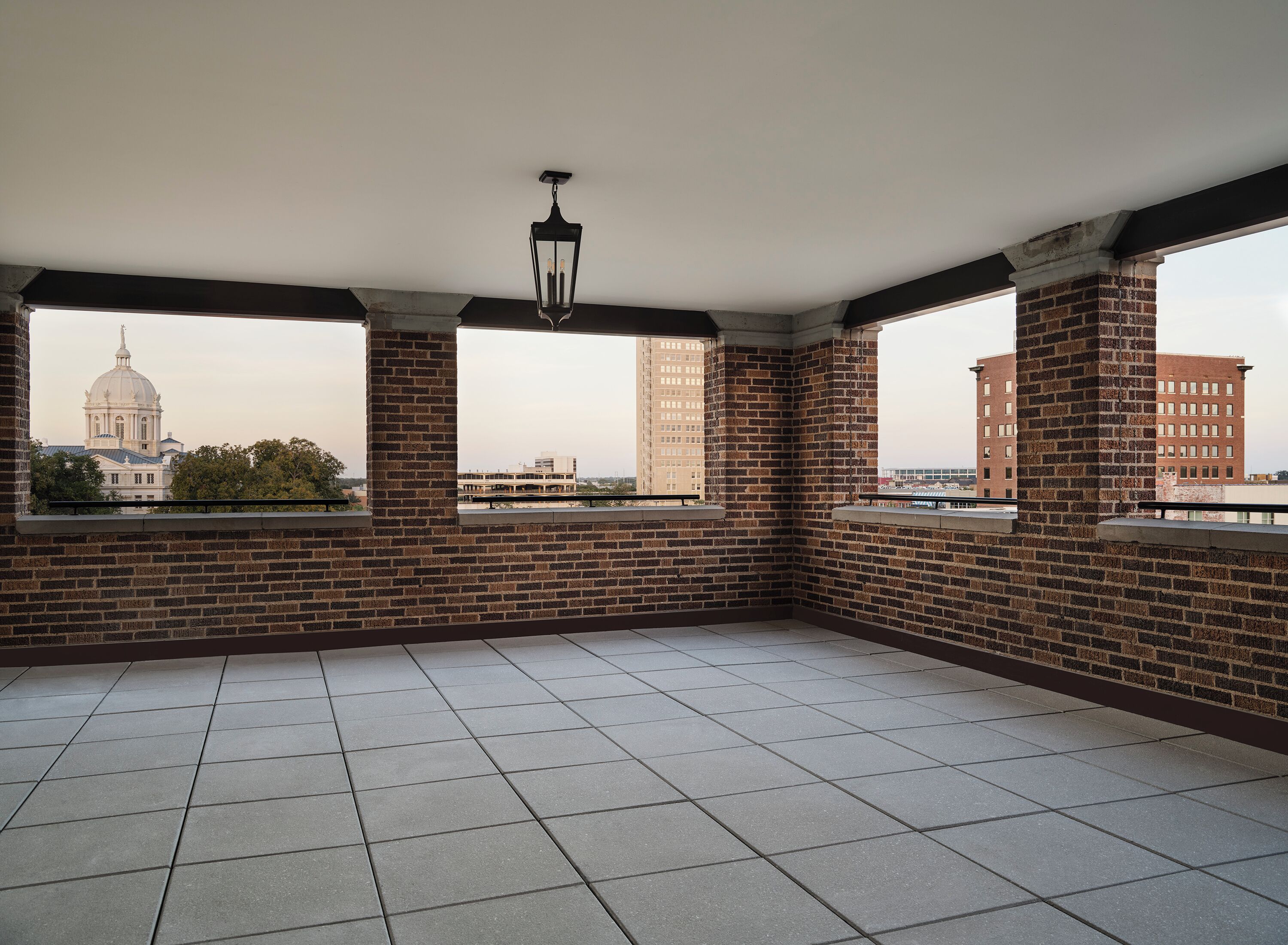 A spacious balcony with tiled flooring, brick walls, and large windows offering views of a cityscape including a prominent building with a dome.