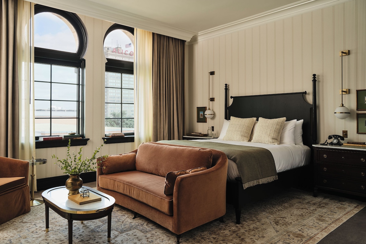 Habitación de hotel con una cama grande, un sofá, una mesita y vistas a la ciudad a través de amplias ventanas.