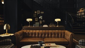 A dark, elegant room with a leather sofa, a round coffee table with books and a bowl, a staircase, and a bookshelf filled with books. - Hotel 1928 (Waco)