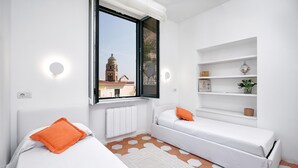 A modern bedroom with two beds, a window with a view of a building, white walls, and a radiator. - Francesca Flat   Amalfi (SA)
