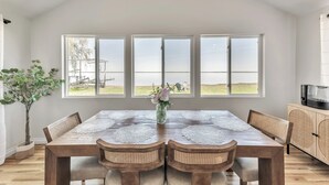 Un coin repas avec une table en bois, quatre chaises et un vase rempli de fleurs. Trois grandes fenêtres se trouvent au-dessus de la table, et une télévision est posée sur un meuble en bois, à droite.