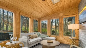 A cozy cabin interior with a sectional sofa, a round coffee table, and large windows offering a view of the surrounding forest. - 6BR Designer Home in Massanutten with Hot Tub, Sauna (McGaheysville)