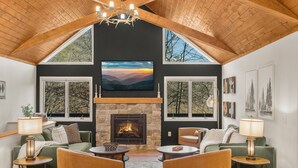 A cozy living room with a stone fireplace, wooden ceiling beams, and large windows offering views of the outdoors. - 6BR Designer Home in Massanutten with Hot Tub, Sauna (McGaheysville)
