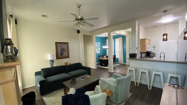 A modern living room with a dark green sofa, a coffee table, and a light blue armchair. There is a dining area with a wooden table and chairs, and a kitchen with a bar counter and stools.