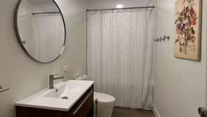 A compact bathroom with a white sink, dark wooden vanity, a round mirror, a white toilet, and a shower curtain. - The North Oak, a Modern Cabin (Washburn)
