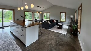 A modern kitchen with a central island, dining area with a sofa, and large windows offering views of greenery. - The North Oak, a Modern Cabin (Washburn)