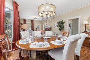Une salle à manger avec une table ronde en bois, dressée pour six personnes, entourée de chaises à revêtement blanc. Un lustre est suspendu au plafond, et une plante en pot est visible à l’arrière-plan.