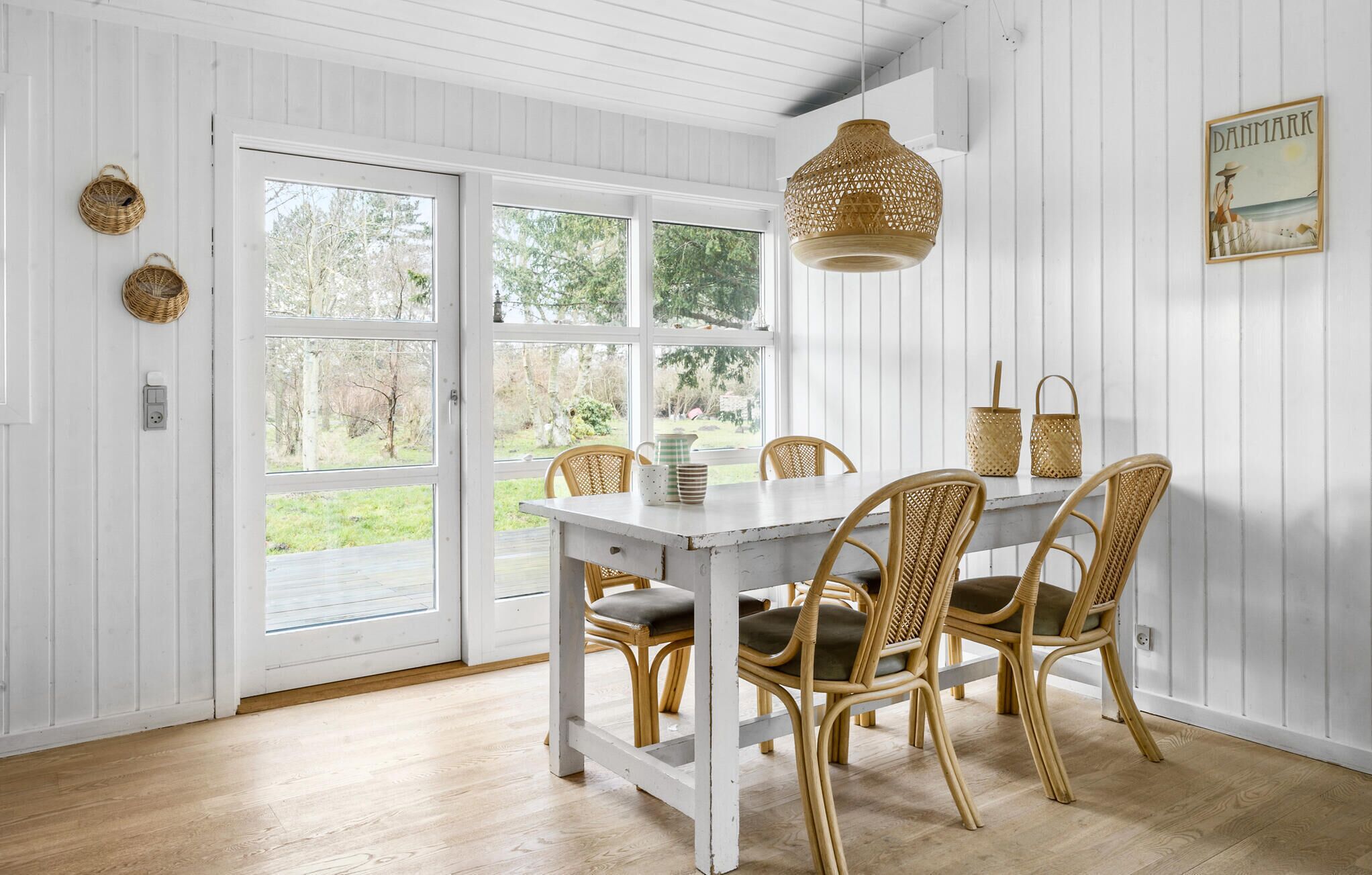 Et spiseområde med et hvidt bord, fletstole, en hængende lampe og et billede i ramme på væggen.