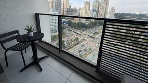 Un balcón con una mesa y silla de metal negro, una planta en maceta y vista a un paisaje urbano con edificios altos y una intersección concurrida.