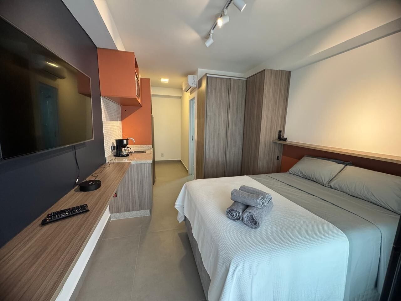 A modern hotel room with a bed, a TV, a desk with a coffee maker, and a bathroom visible in the background.