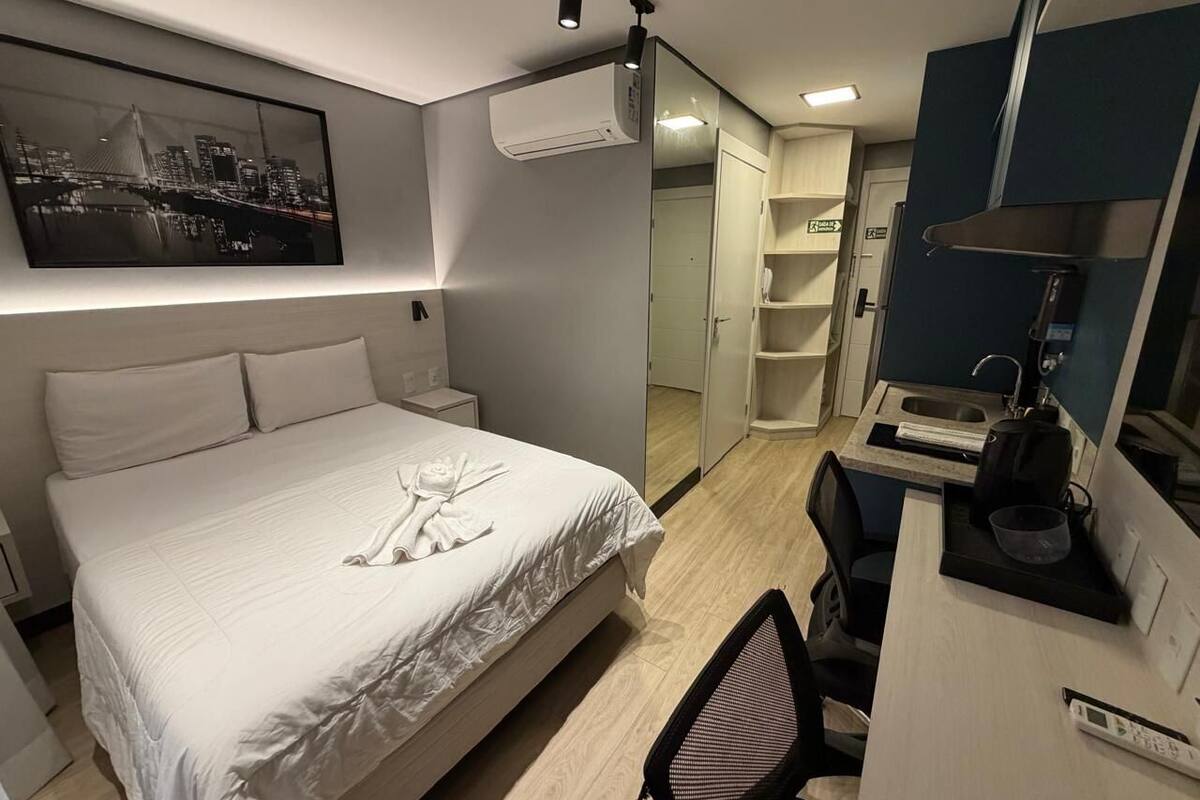 A hotel room with a bed, a desk, a chair, a mirror, and a wall-mounted air conditioner.
