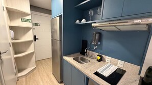 Una cocina compacta con un refrigerador de acero inoxidable, un fregadero y una cafetera. Hay estantes sobre la encimera y una puerta con un cartel que indica la salida.