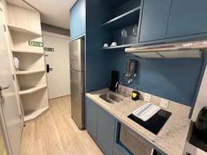 Une cuisine compacte équipée d’un réfrigérateur en acier inoxydable, d’un évier et d’une machine à café. Des étagères se trouvent au-dessus du plan de travail, et une porte, agrémentée d’une signalétique, indique la sortie.