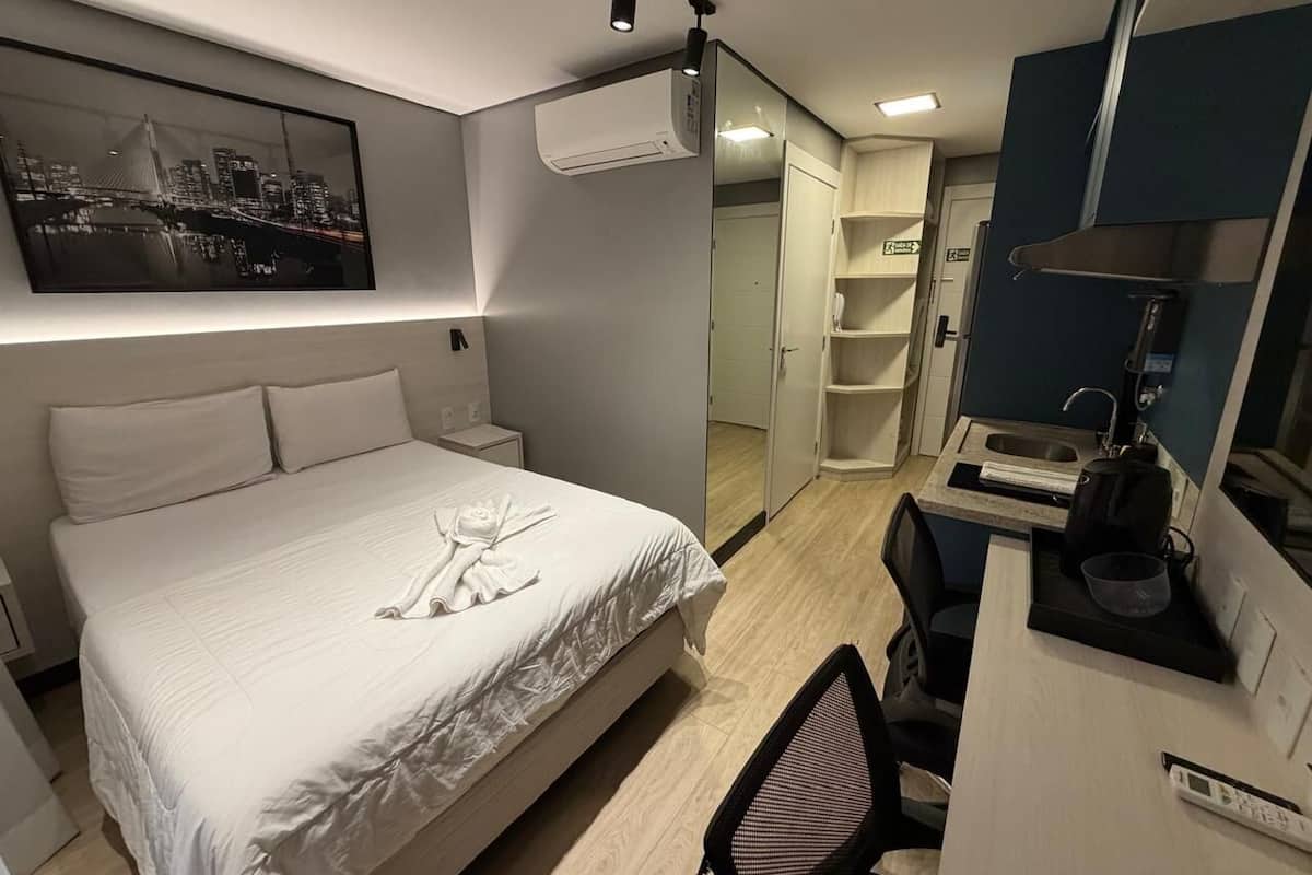 Quarto de hotel com cama, escrivaninha, cadeira, espelho e ar-condicionado fixado na parede.