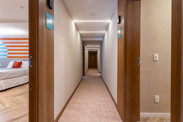 A hotel corridor with wooden doors, a carpeted floor, and a bed with a red pillow in the background.