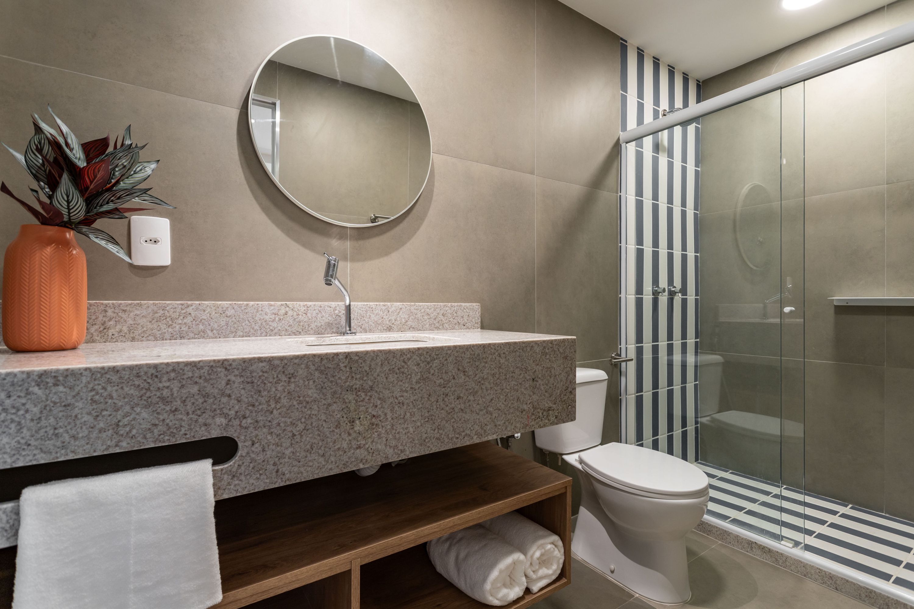 A modern bathroom with a granite countertop, a round mirror, a toilet, and a glass shower enclosure.