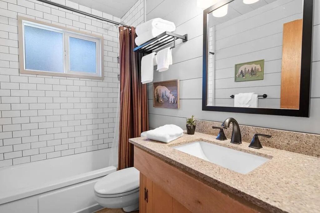 Een badkamer met een witte betegelde muur, een raam met zonwering, een douche met gordijn, een wastafel met spiegel, en een toilet.