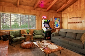 Un salon chaleureux avec un décor intérieur en bois, un canapé vert, une table basse avec un jeu de société et une borne d’arcade rétro.